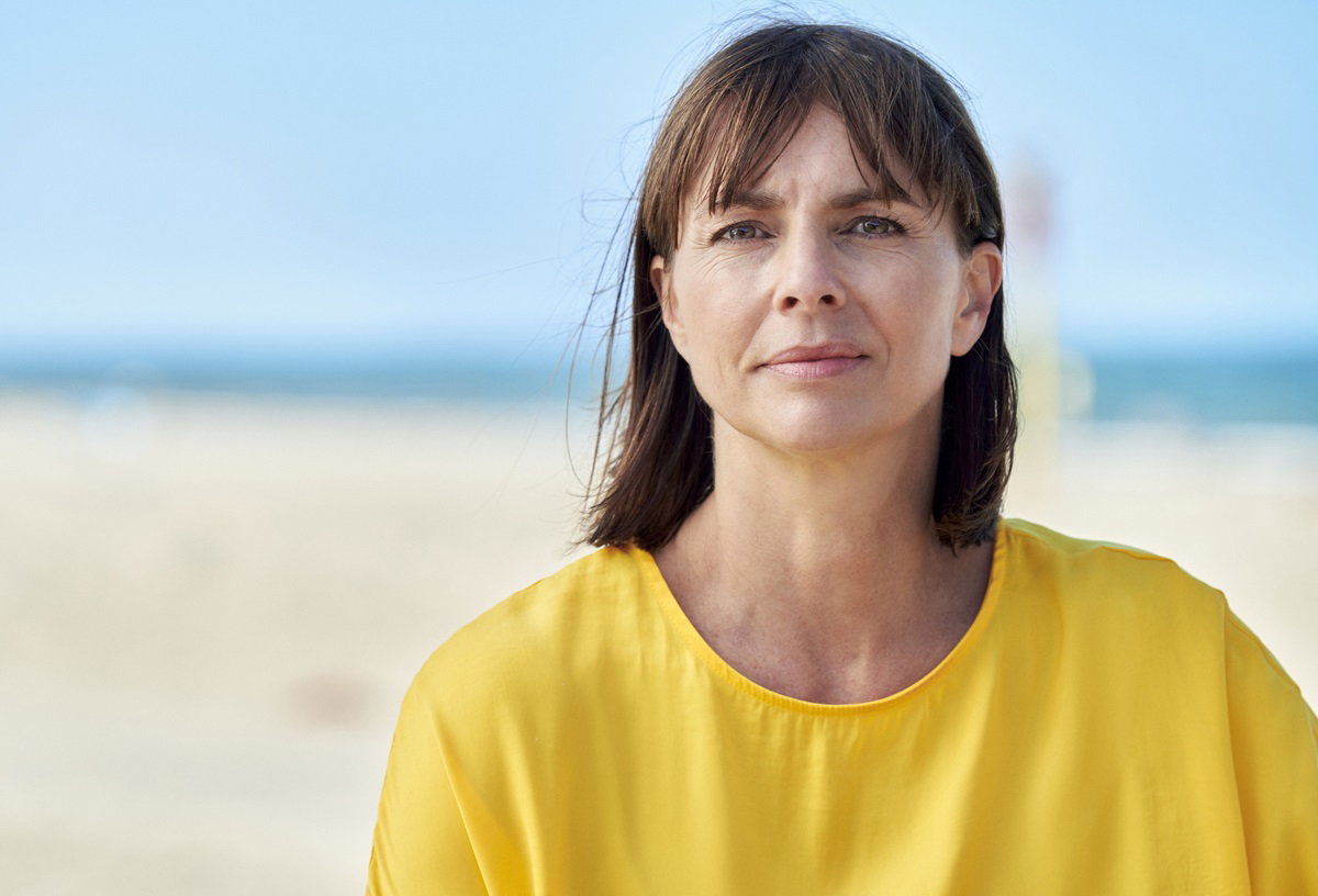 kobieta w średnim  wieku około 40+ w żółtej bluzce siedzi na tle plaży i morza z poważną miną