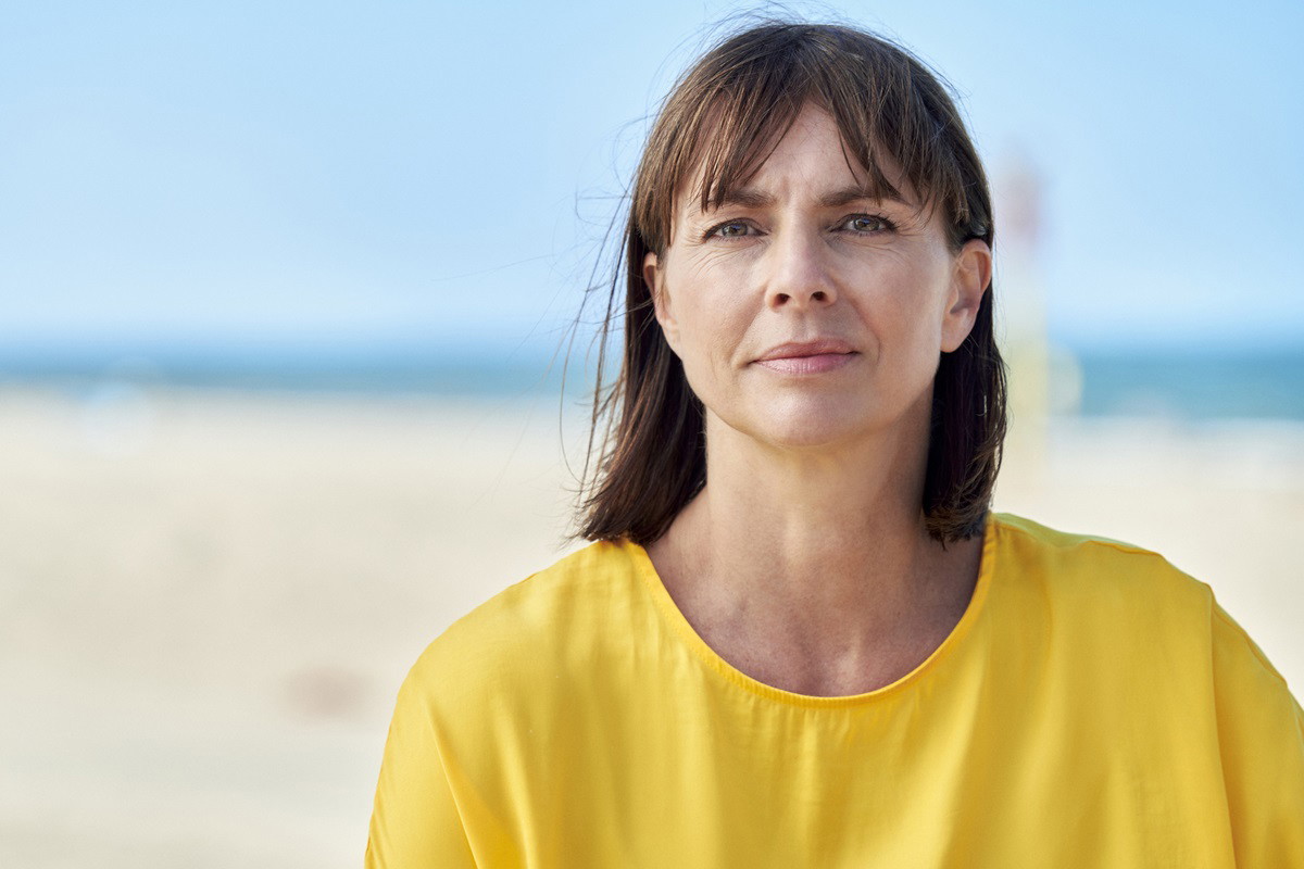kobieta w średnim wieku około 40+ w żółtej bluzce siedzi na tle plaży i morza z poważną miną