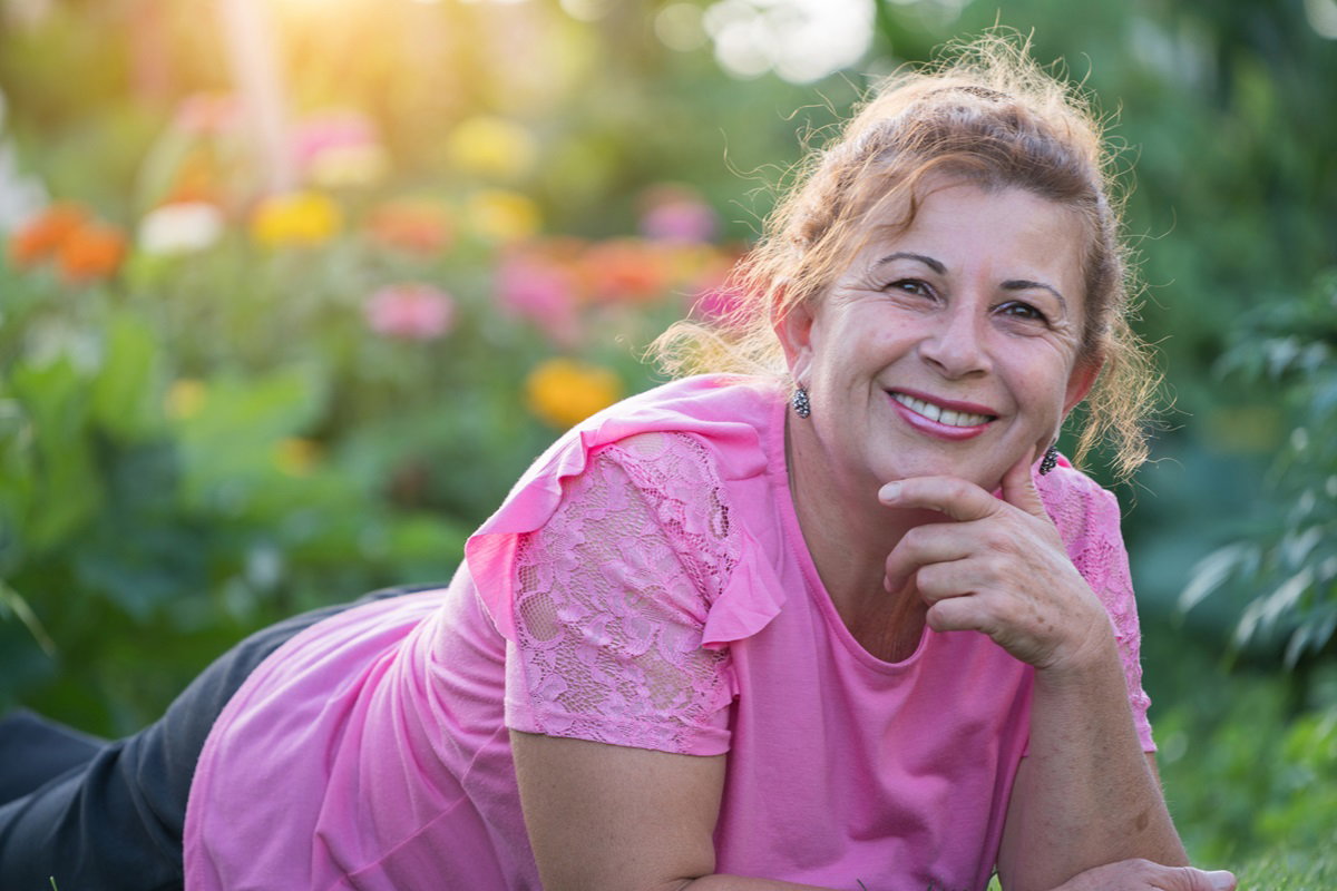 kobieta w wieku około 60 plus leży na trawie na działce w różowej bluzce i się uśmiecha