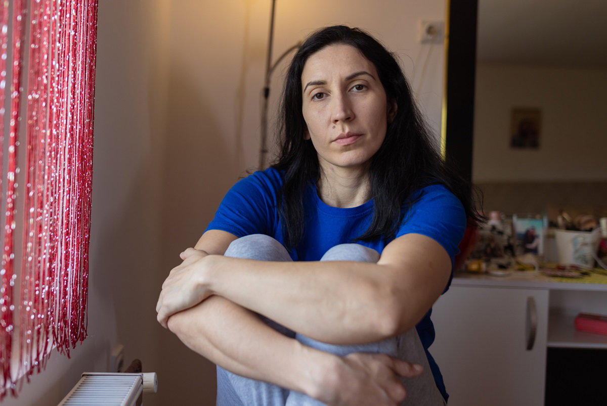 kobieta w wieku około 40 lat z ciemnymi długimi włosami siedzi w pokoju w niebieskiej bluzce i trzyma kolana ze smutną miną