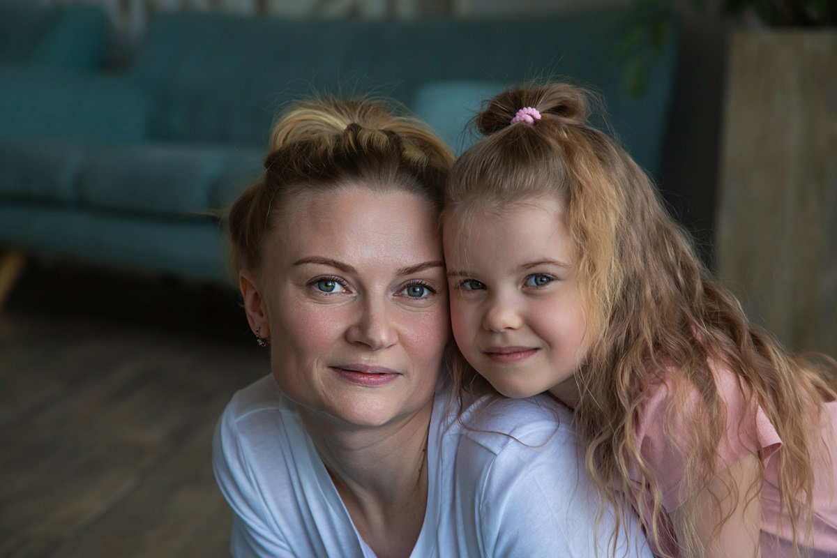 kobieta w średnim wieku siedzi na podłodze z córeczką z długimi włosami i lekko się uśmiechają