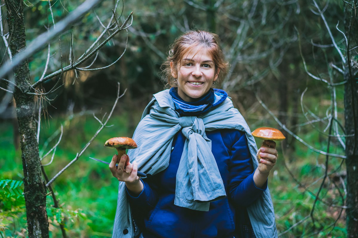 młoda kobieta stoi w lesie i trzyma dwa grzyby w rękach uśmiechając się