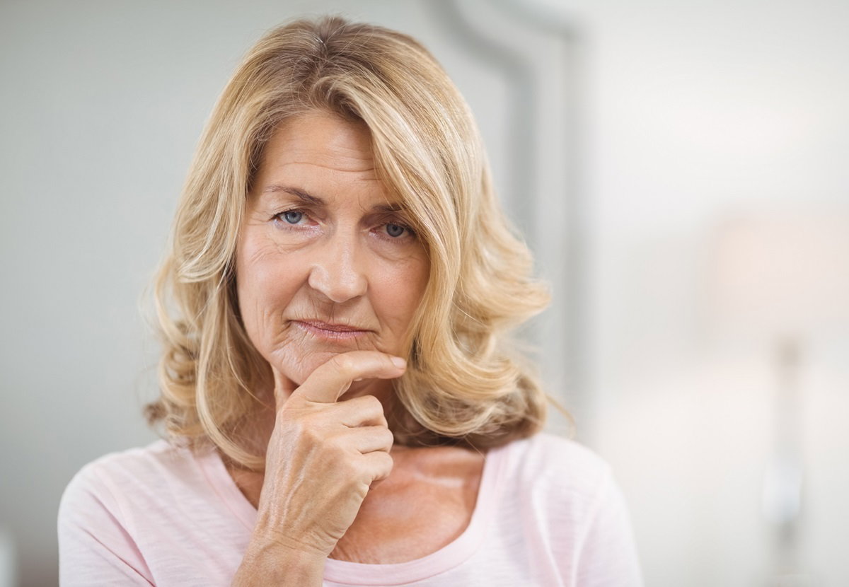 kobieta w wieku około 60+ blondynka stoi na tle białego wnętrza i patrzy smutnym wzrokiem