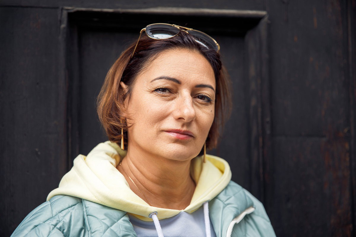 kobieta w średnim wieku na tle ciemnych drzwi stoi i patrzy powątpiewającym wzrokiem