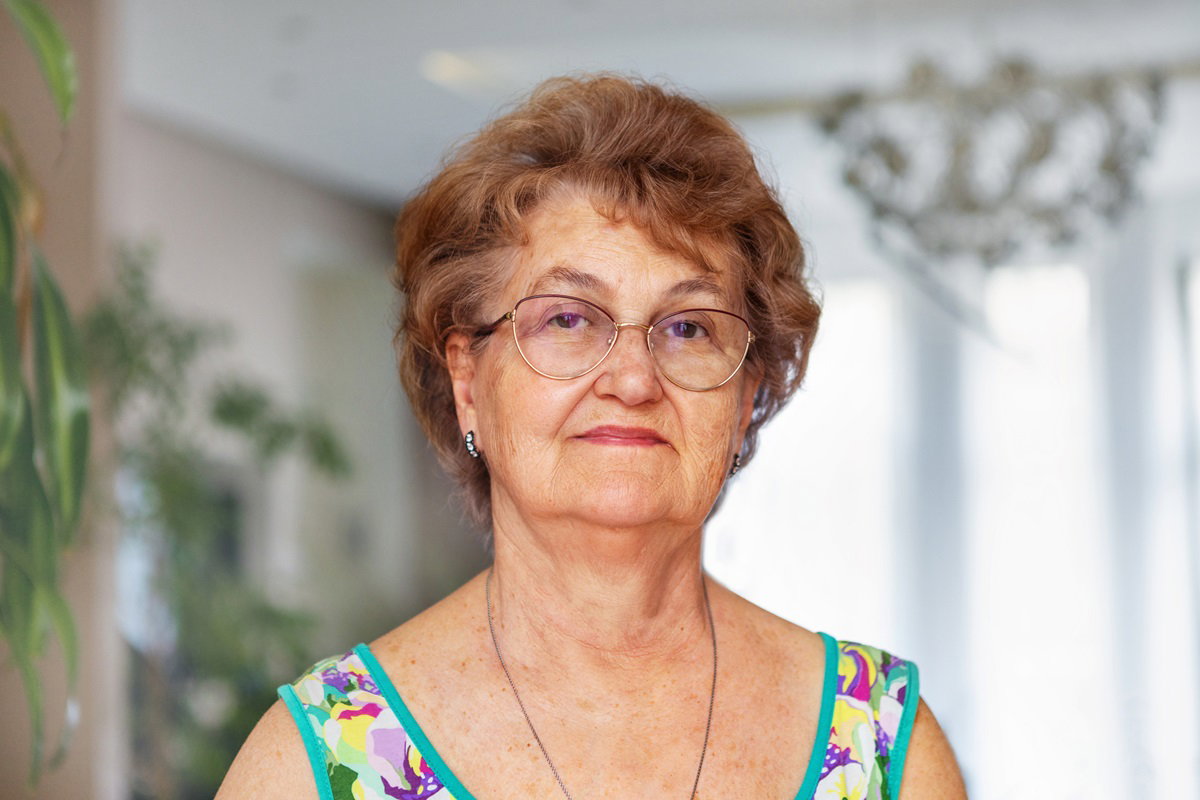 kobieta seniorka w wieku około 65+ w okularach i sukience bez rękawów stoi na tle salonu z poważną i lekko zdegustowaną miną