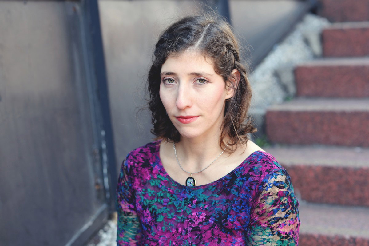 młoda kobieta siedzi na schodach w kolorowej bluzce i z naszyjnikiem ma zawiedzioną minę