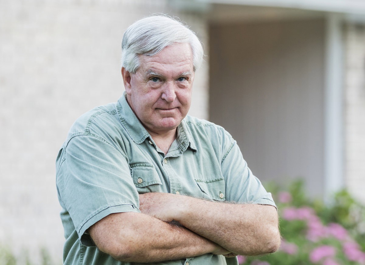 mężczyzna w wieku senioralnym z siwymi włosami stoi w ogrodzie przed domem z założonymi rękami i zdenerwowana miną