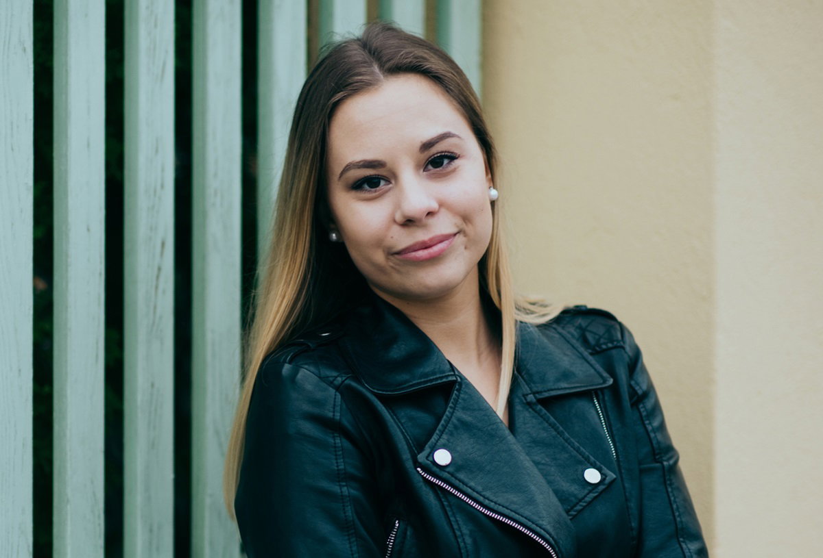 młoda kobieta blondynka w kurtce ramonesce stoi na tle ściany z niepewnym uśmiechem