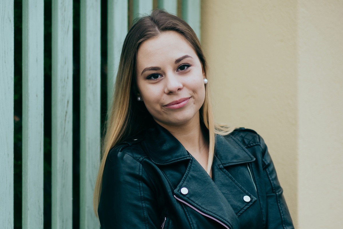 młoda kobieta blondynka w kurtce ramonesce stoi na tle ściany z niepewnym uśmiechem