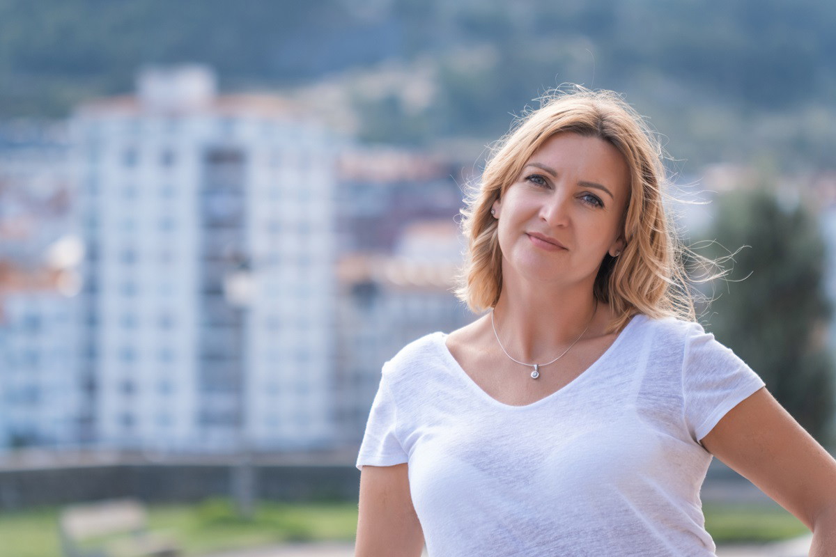 kobieta w średnim wieku blondynka stoi na tle osiedla z lekkim uśmiechem