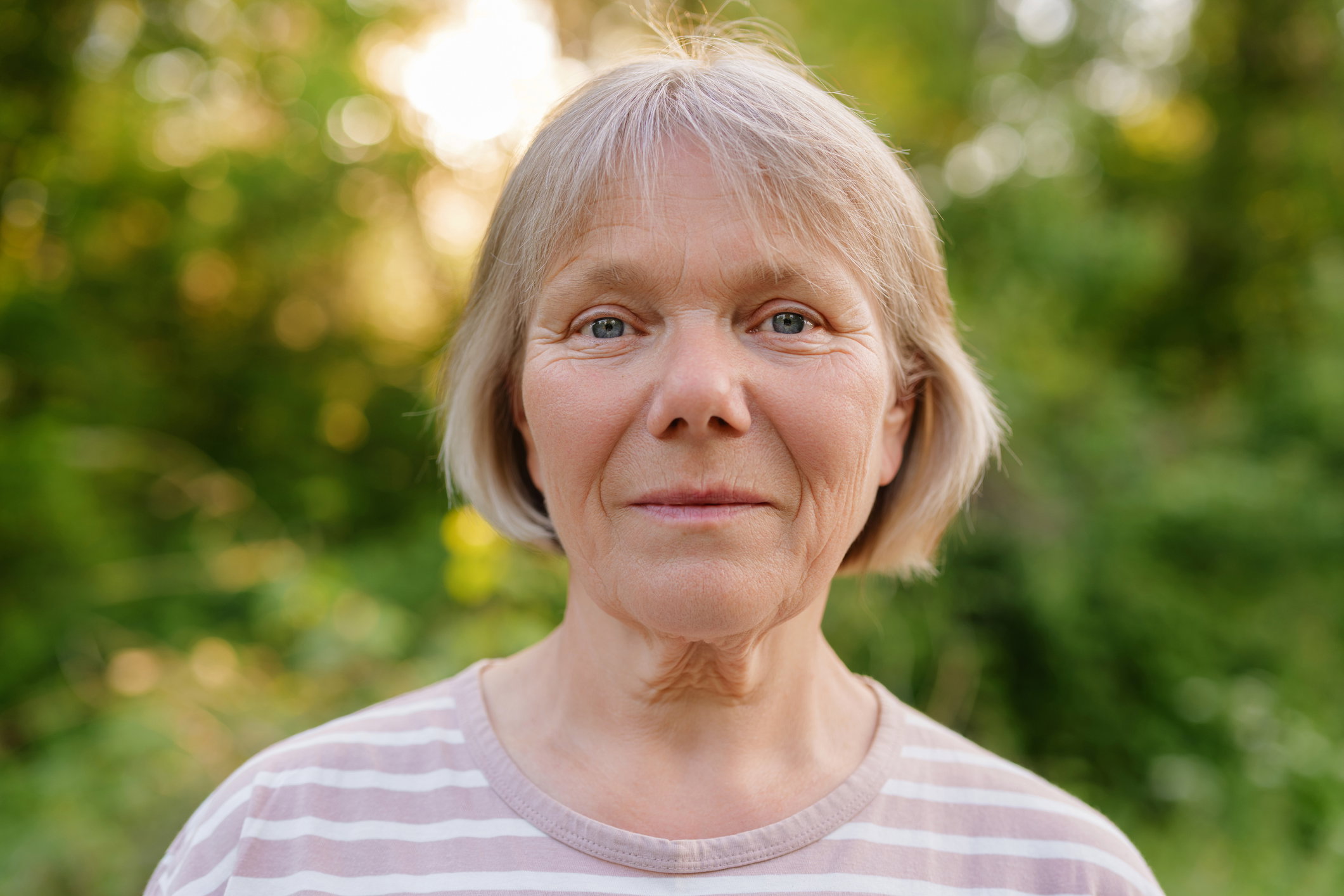 A clear, direct outdoor portrait of a serene middle-aged woman with short, light hair and blue eyes, gazing straight at the camera against a blurred, sunlit green background.