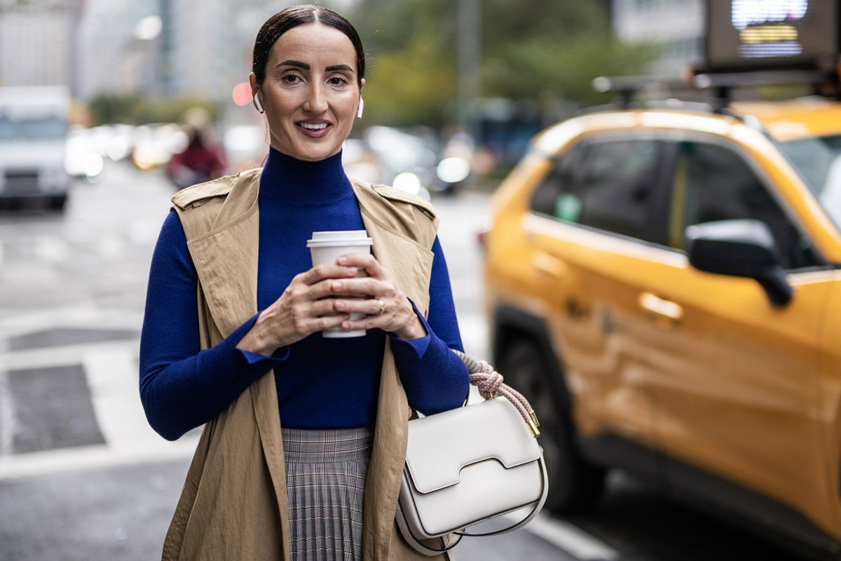 kobieta w średnim wieku w płaszczu bez rękawów i kubkiem kawy w reku stoi na ulicy na tle żółtej taksówki