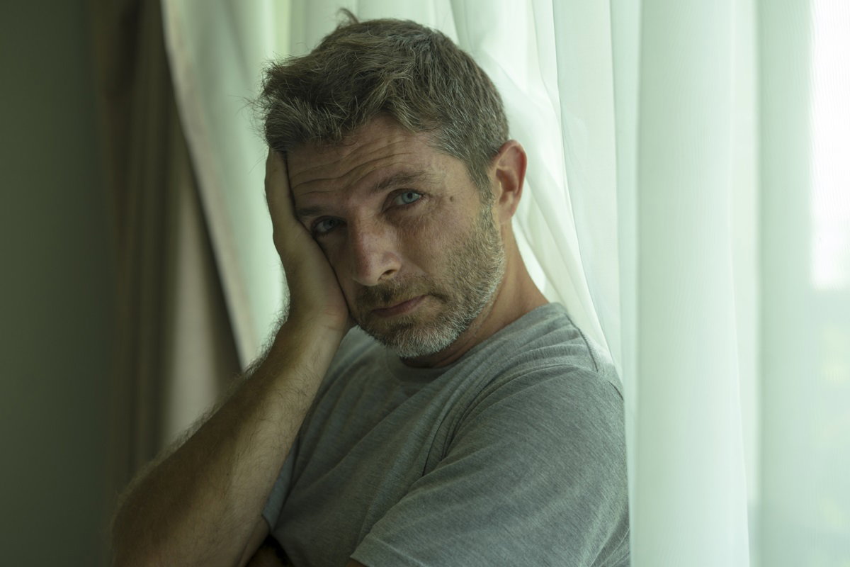 mężczyzna w średnim wieku z zarostem stoi na tle okna zrozpaczony