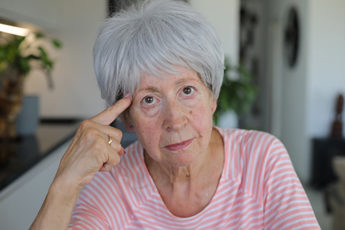 kobieta seniorka z krótkimi siwymi włosami siedzi w kuchni i wskazuje palcem na głowę