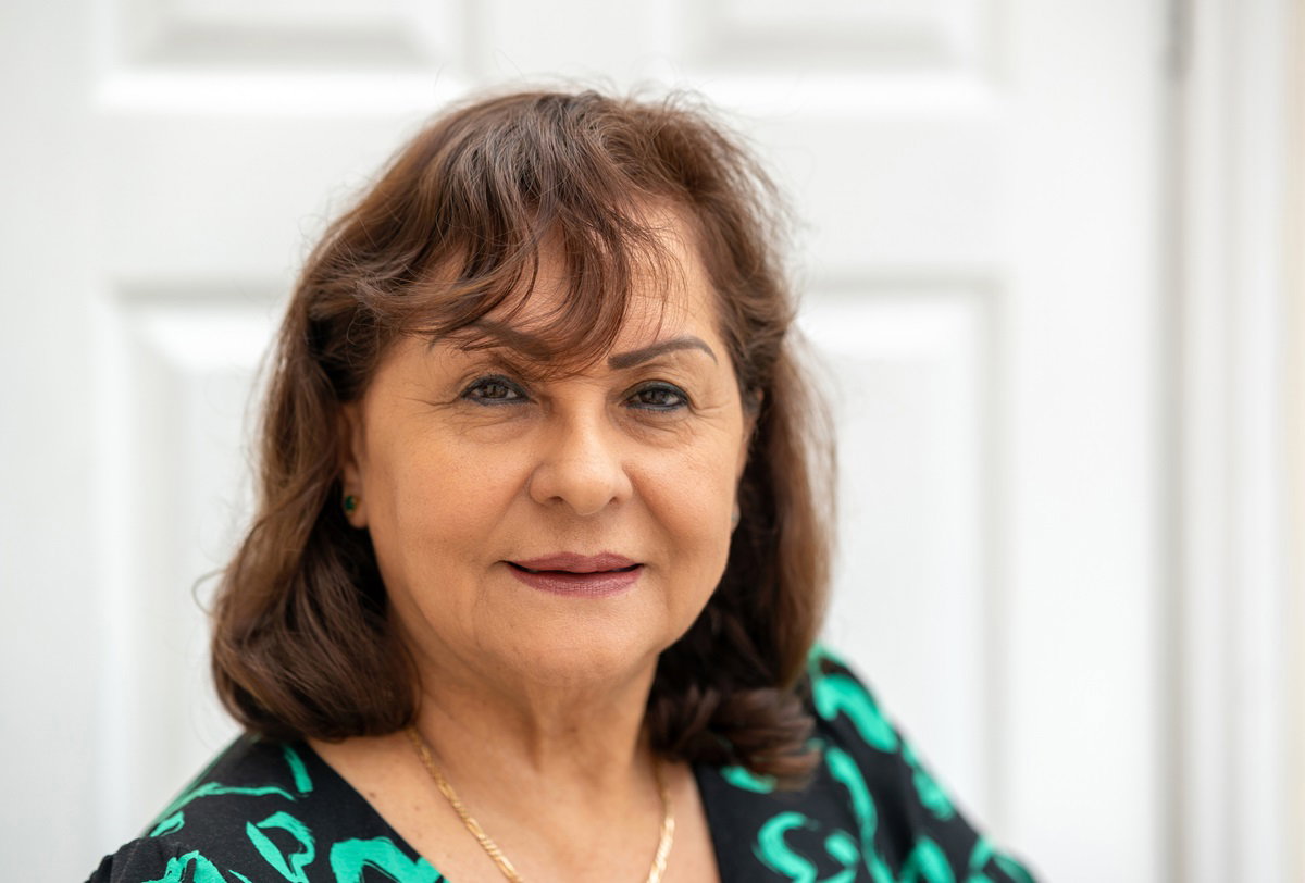 kobieta w wieku około 60 lat w sukience w zielone wzory stoi na tle drzwi i patrzy z lekkim uśmieszkiem na twarzy