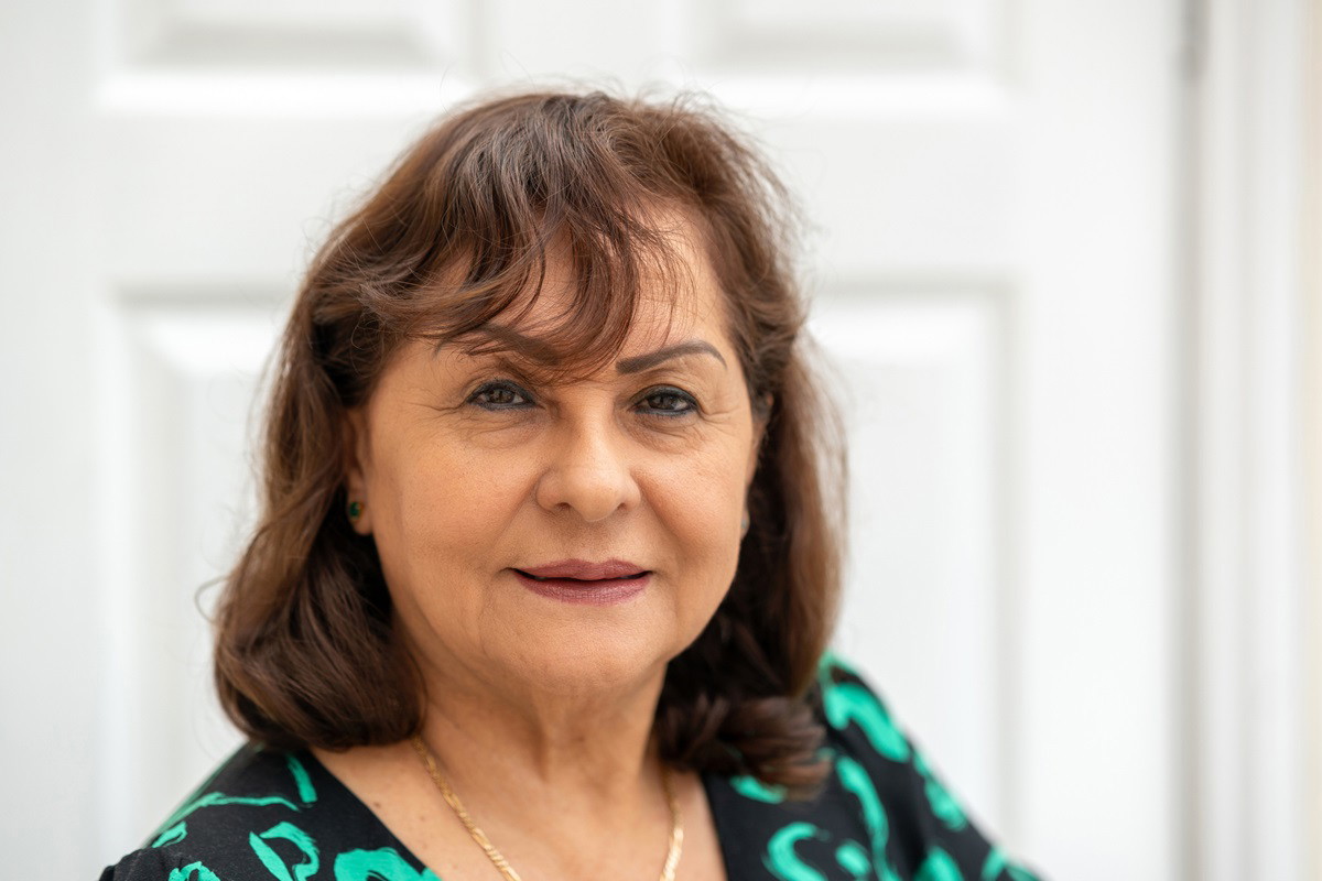 kobieta w wieku około 60 lat w sukience w zielone wzory stoi na tle drzwi i patrzy z lekkim uśmieszkiem na twarzy