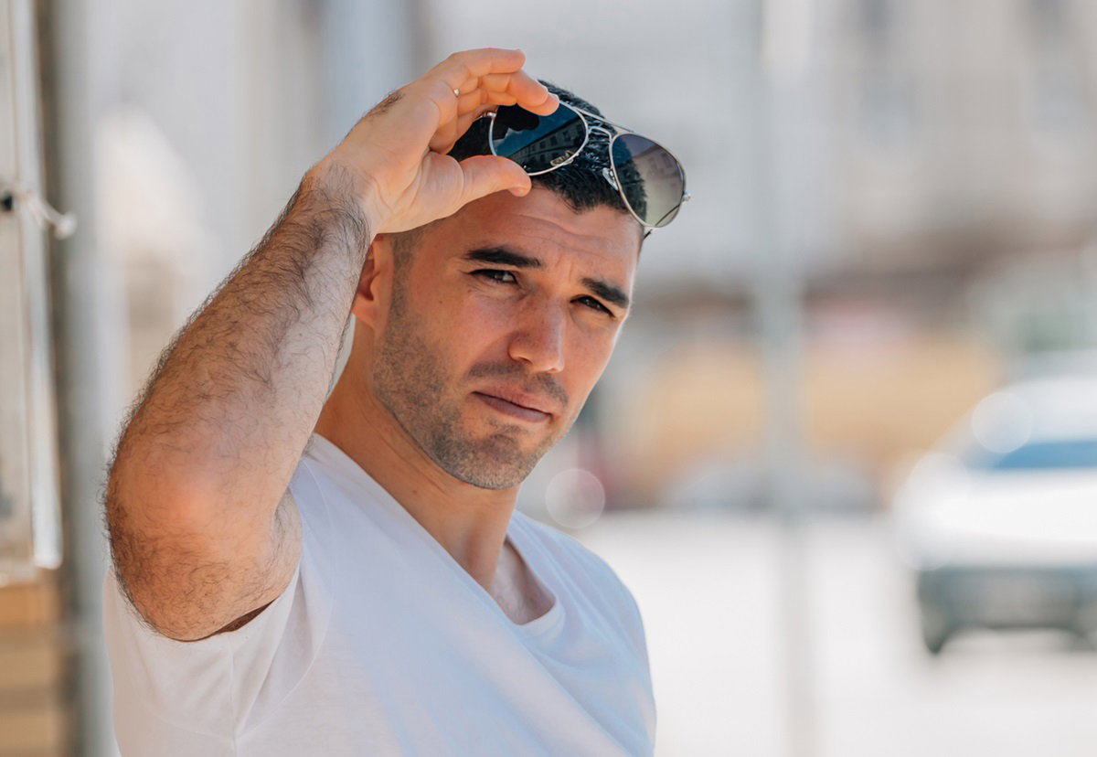 młody mężczyzna stoi na ulicy w słońcu ze zmartwioną min i trzyma okulary na głowie