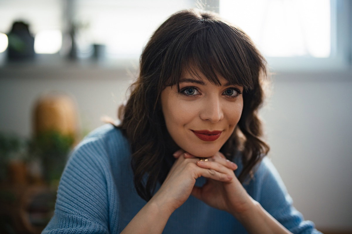 kobieta w wieku około 30 plus w błękitnym swetrze siedzi z rękami pod brodą i lekko się uśmiecha