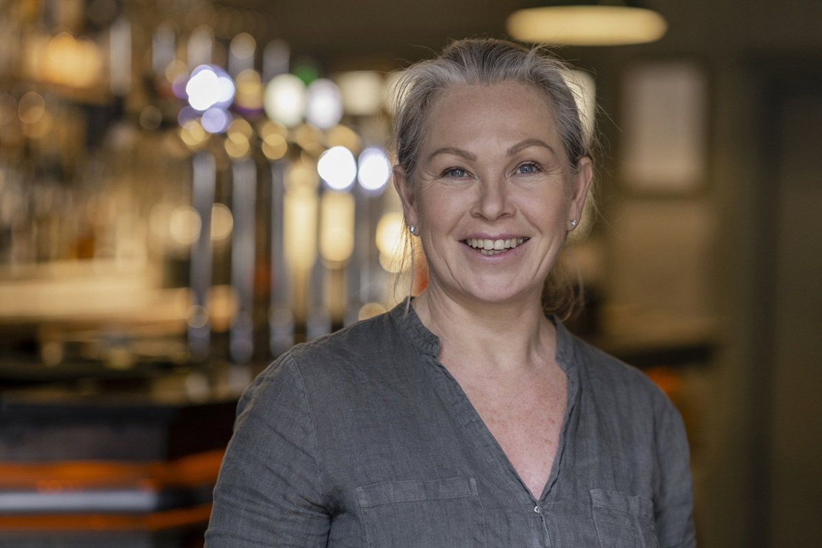 kobieta w wieku 50 plus w szarej koszuli stoi uśmiechnięta w swojej cukierni