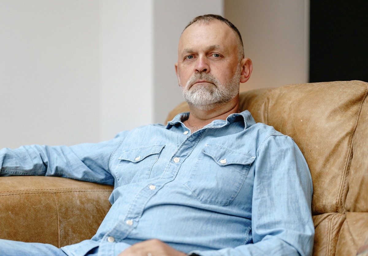 mężczyzna 65 plus z zarostem w jeansach i jeansowej koszuli siedzi na kanapie zamyślony