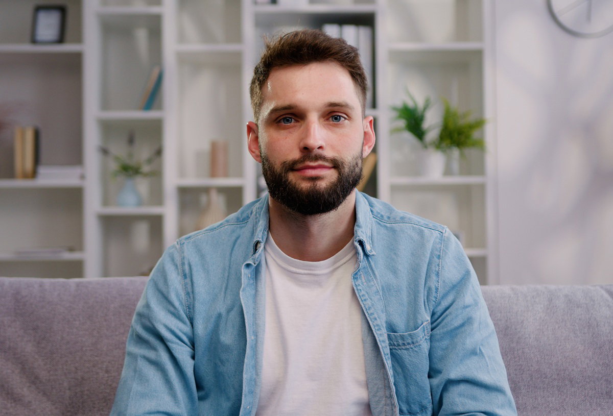 mężczyzna w wieku około 35 lat z zarostem siedzi na kanapie i patrzy poważnym wzrokiem