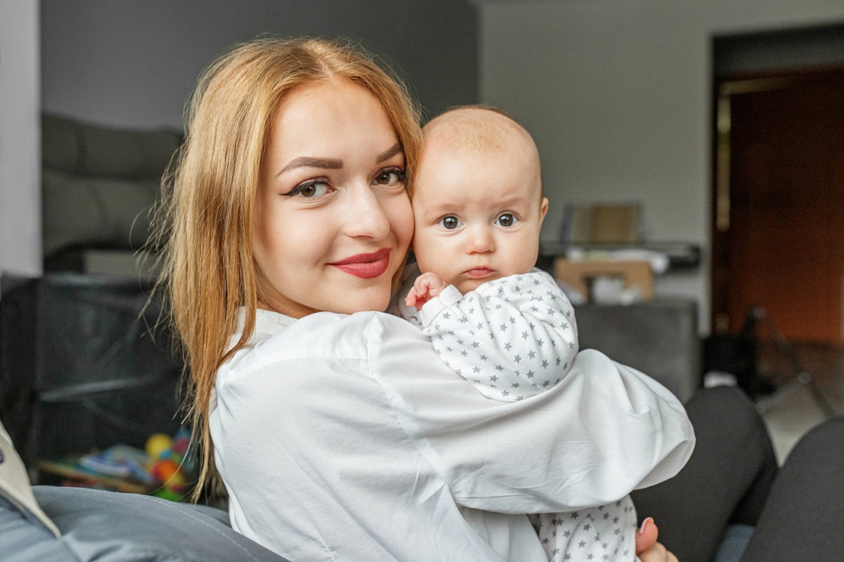 młoda kobieta 30 plus z uroczym dzieckiem na rękach stoi w salonie i uśmiecha się