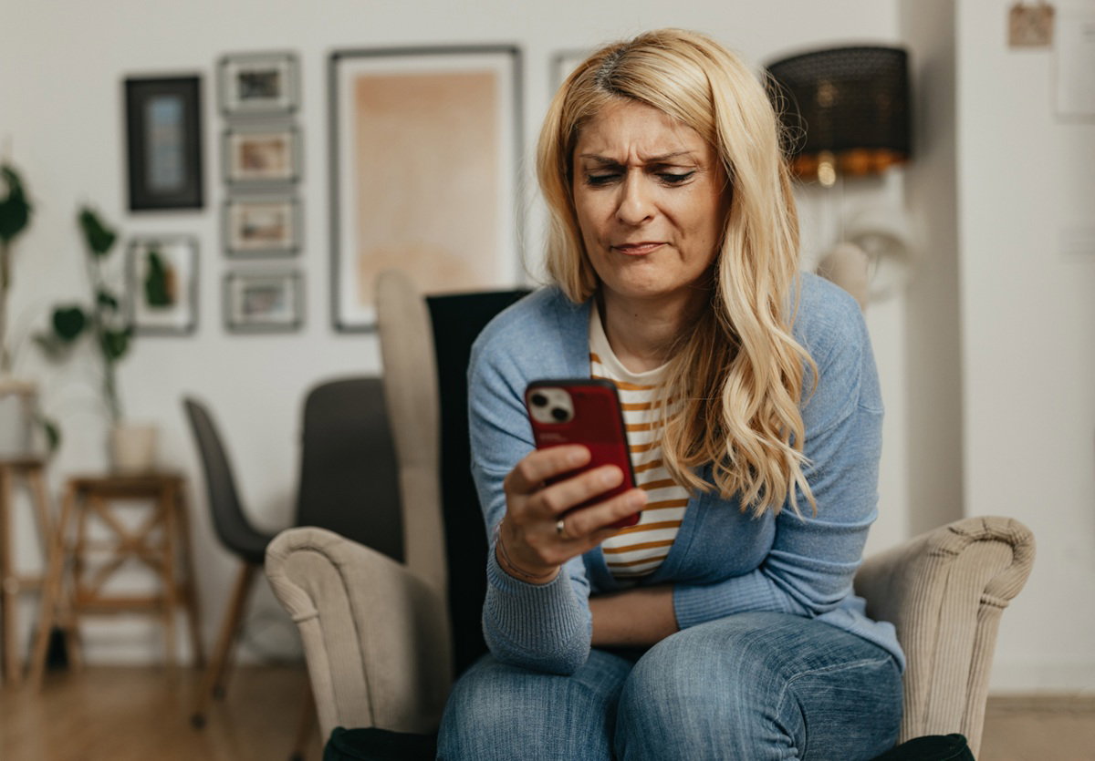 kobieta w wieku 35 plus blondynka siedzi na fotelu i patrzy w telefon z niesmakiem