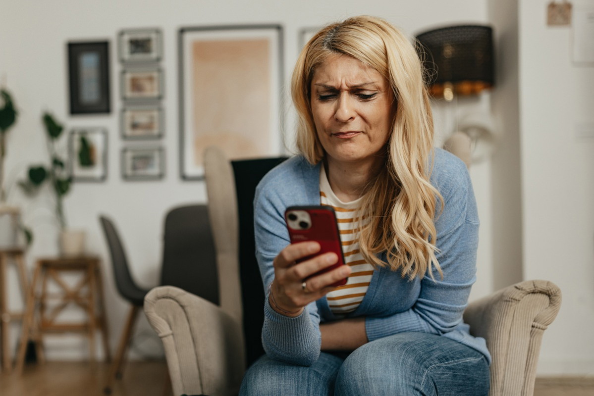 kobieta w wieku 35 plus blondynka siedzi na fotelu i patrzy w telefon z niesmakiem