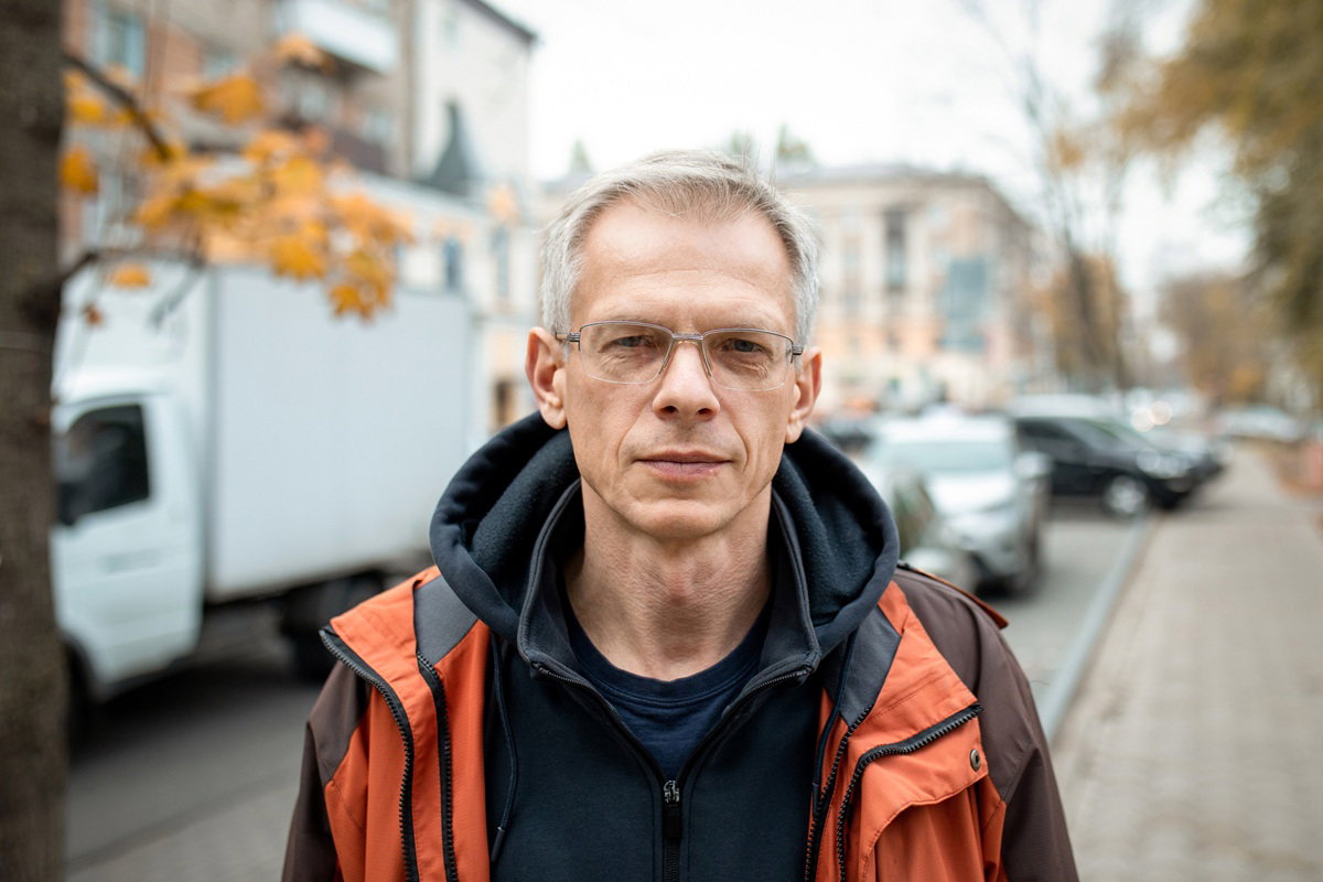 mężczyzna 45 plus w okularach siwiejący w kurtce stoi na ulicy i patrzy poważnym wzrokiem