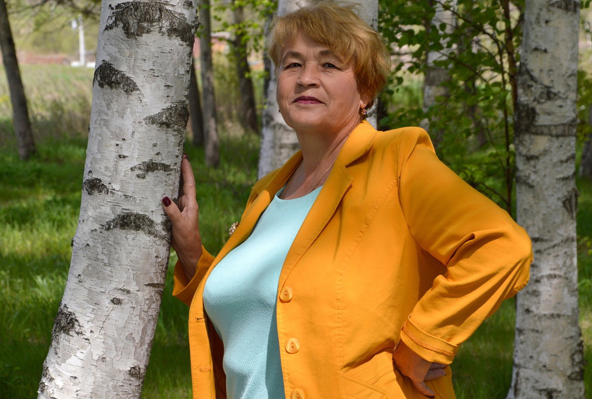 kobieta w wieku 65 plus w żółtej marynarce stoi przy brzozach zamyślona