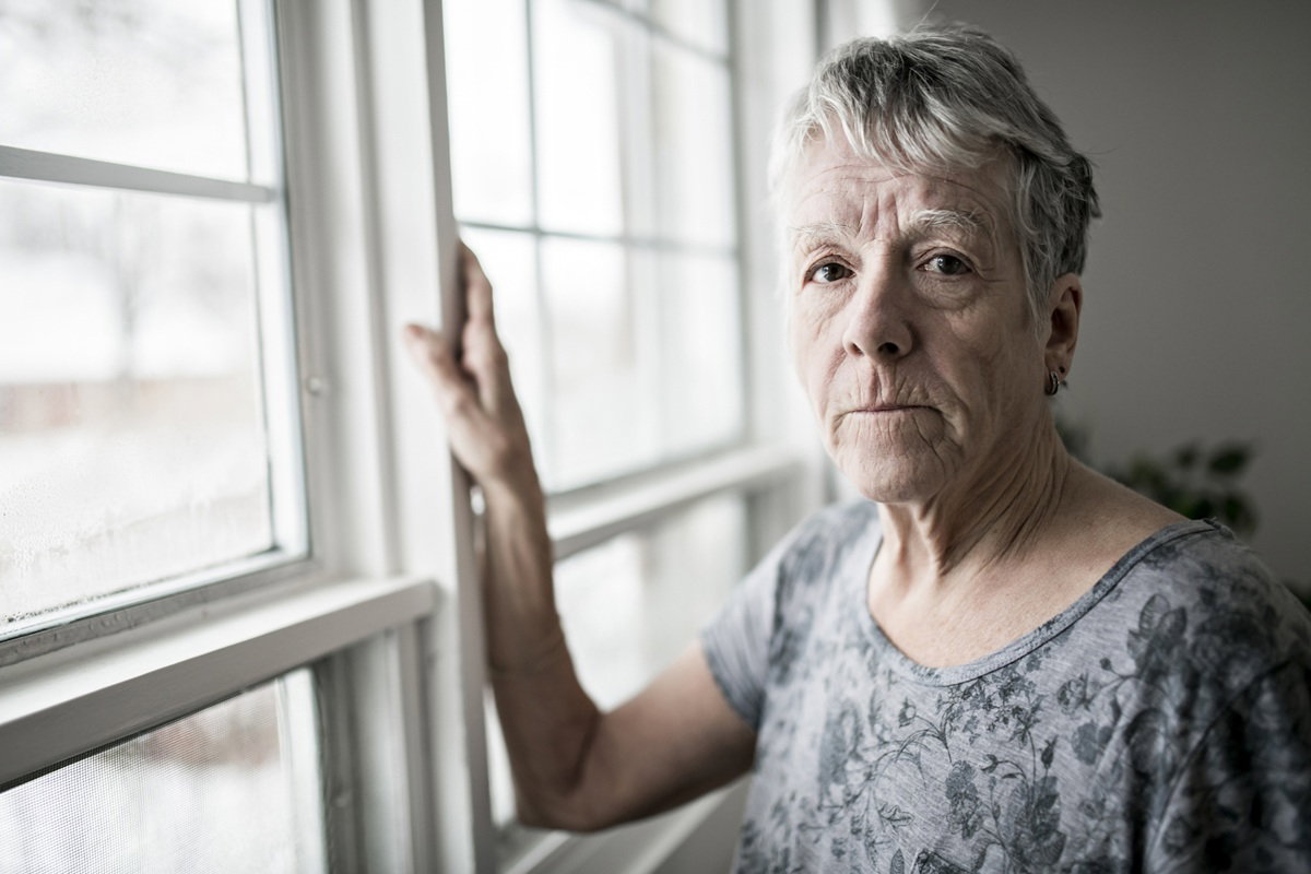kobieta w wieku około 70 plus siwe krótkie włosy stoi przy oknie i ma bardzo smutny wyraz twarzy