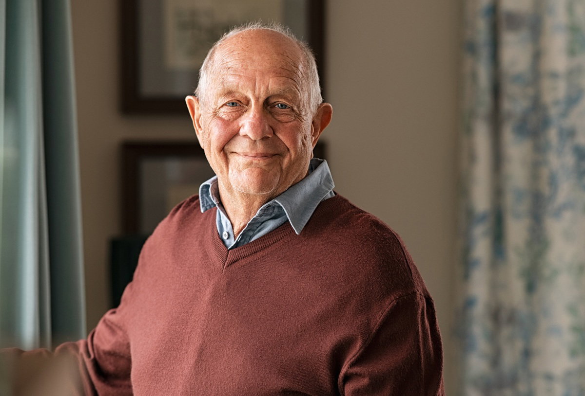 mężczyzna senior w wieku około 70 plus w brązowym swetrze stoi w salonie i się uśmiecha