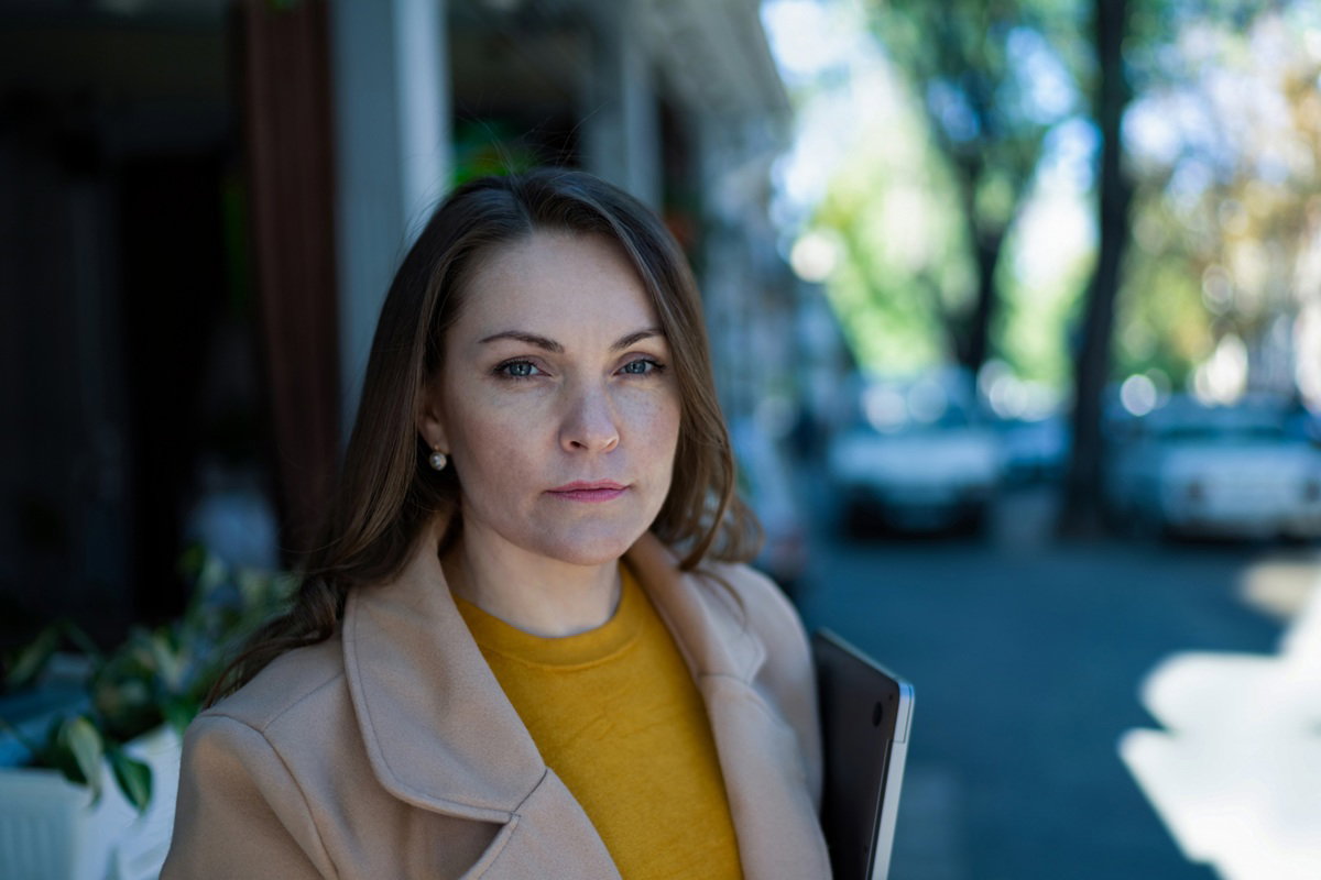 kobieta w wieku 35 plus w płaszczu i z laptopem pod pachą stoi na zewnątrz i patrzy poważnym wzrokiem