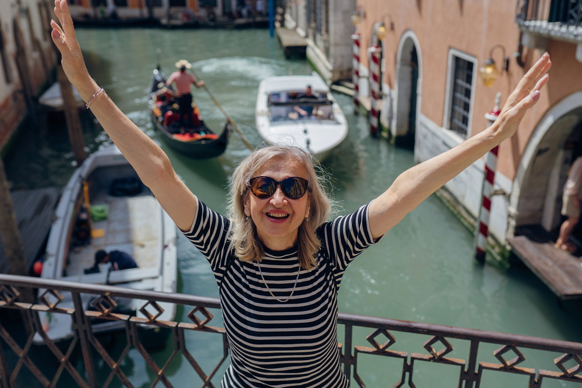 kobieta w wieku 60 plus w okularach stoi z rękami do góry na mostku nad kanałem w Wenecji