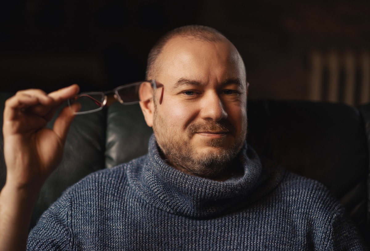 mężczyzna w wieku 40 plus w szarym swetrze siedzi zamyślony z okularami w ręku