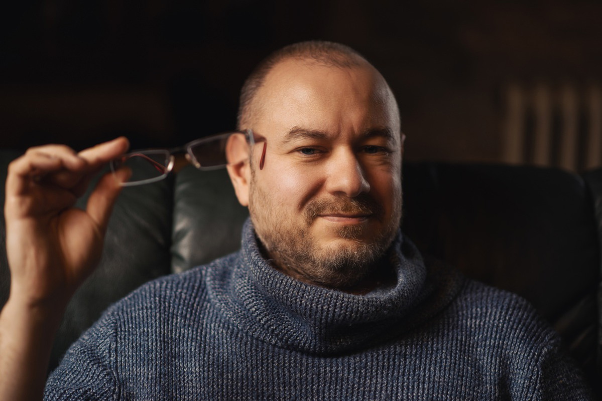 mężczyzna w wieku 40 plus w szarym swetrze siedzi zamyślony z okularami w ręku