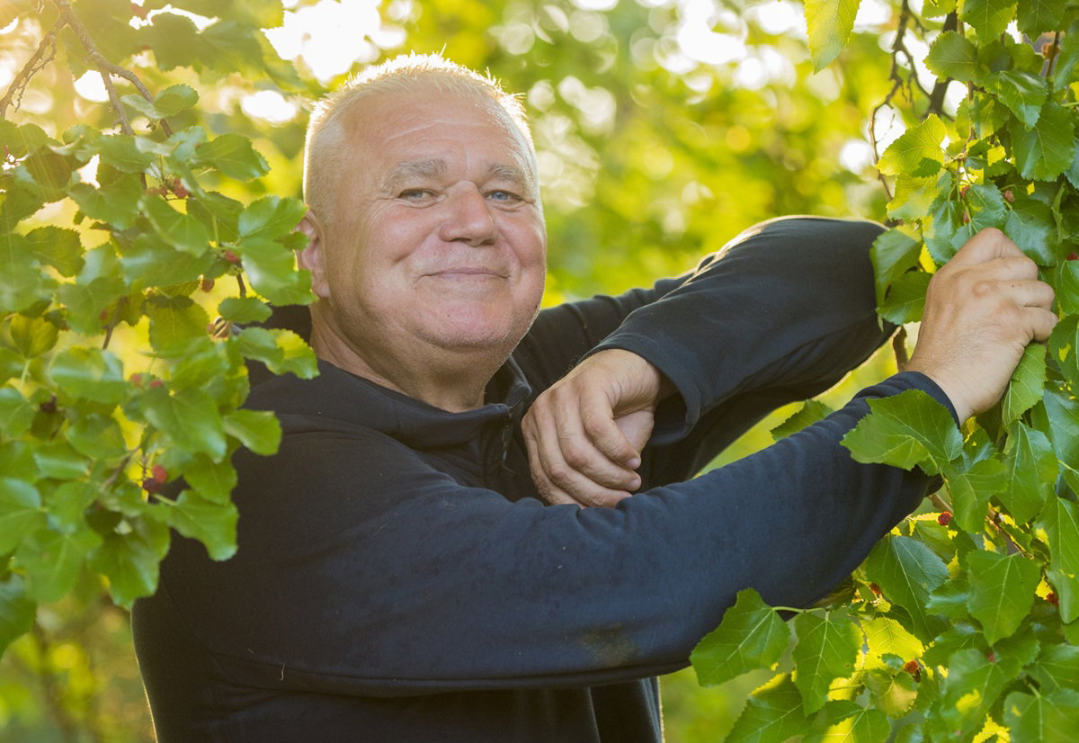 mężczyzna w wieku około 60 lat w czarnej bluzie uśmiecha się w krzakach