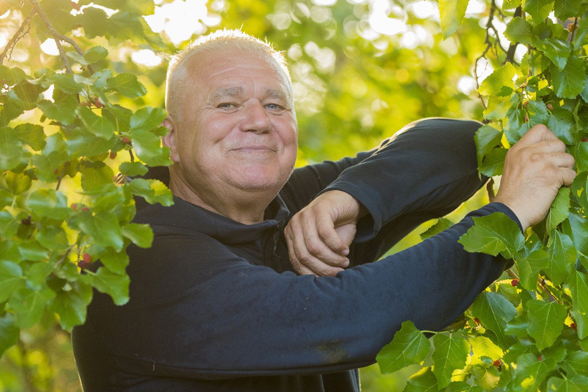 mężczyzna w wieku około 60 lat w czarnej bluzie uśmiecha się w krzakach