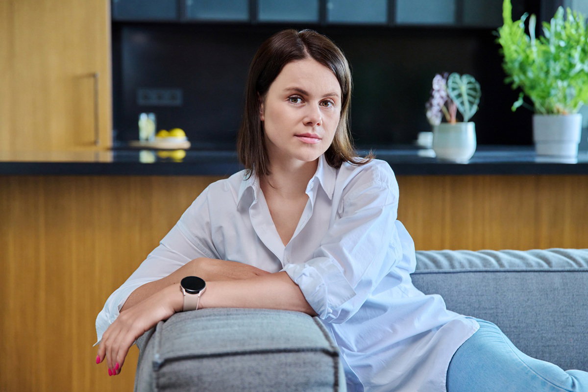 kobieta 30 plus w białej koszuli siedzi na tle kuchni na kanapie zamyślona