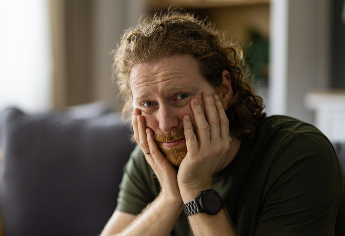 mężczyzna w wieku około 35 lat z włosami i zarostem w kolorze rudym siedzi zmartwiony na kanapie