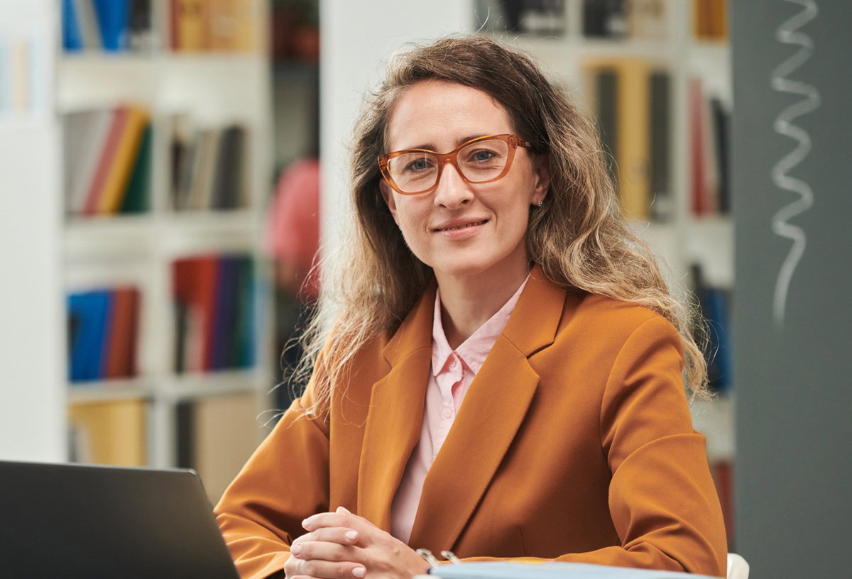 kobieta w wieku około 35 plus w pomarańczowej marynarce i w okularach siedzi przy biurku na tle książek i tablicy