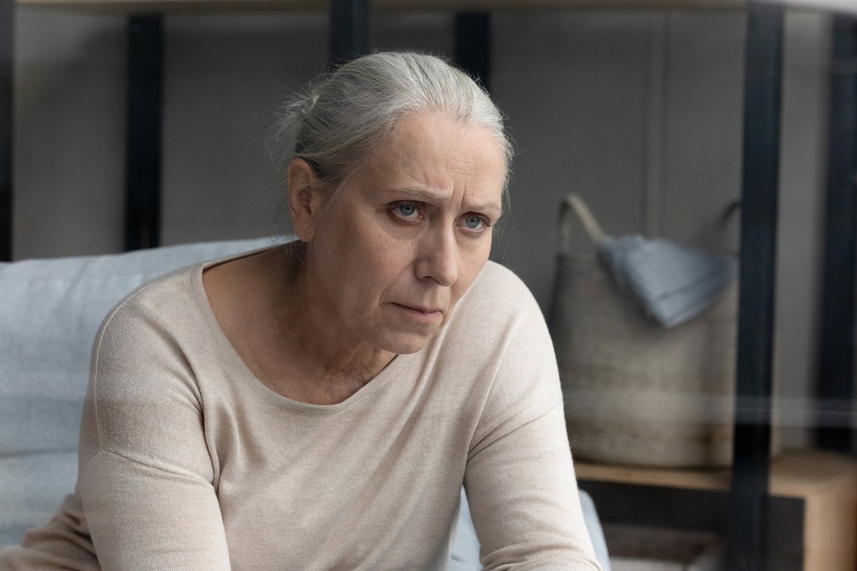 kobieta w wieku 60 plus z siwymi włosami siedzi i patrzy smutno w jeden punkt