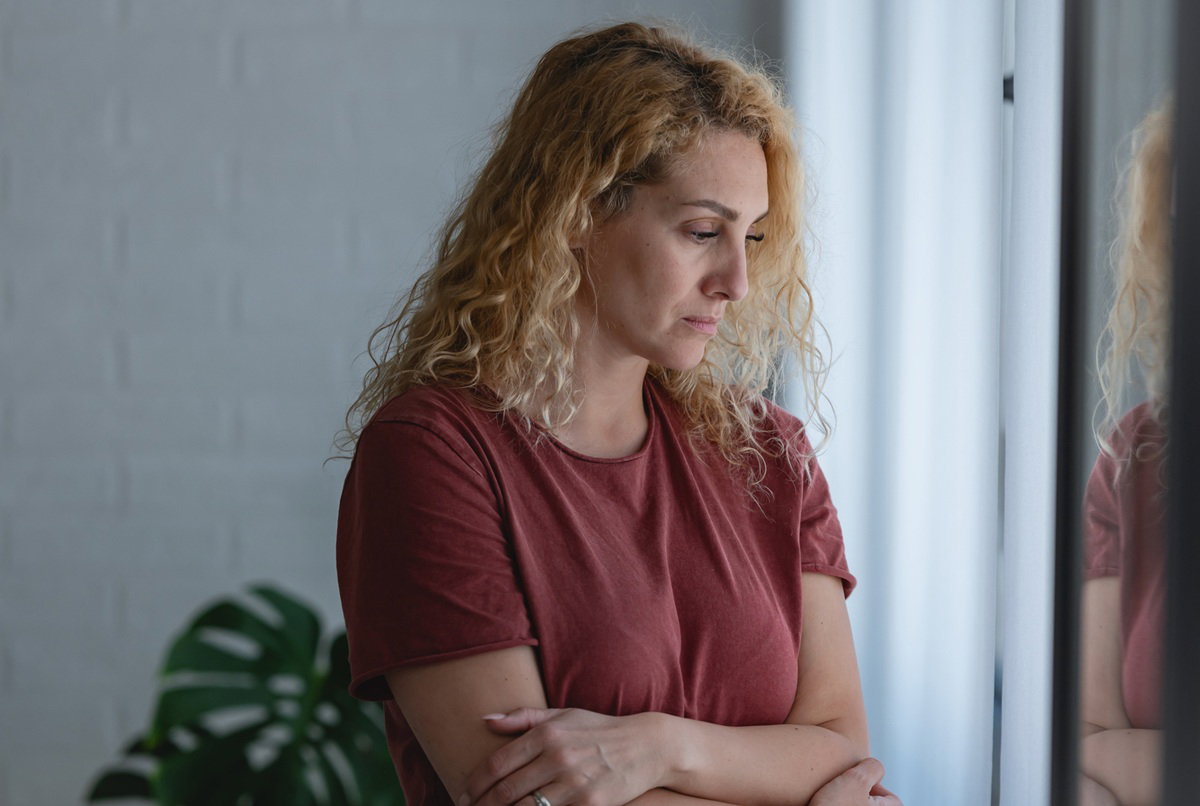 kobieta w wieku 35 plus blondynka kręcone włosy stoi przy oknie smutna