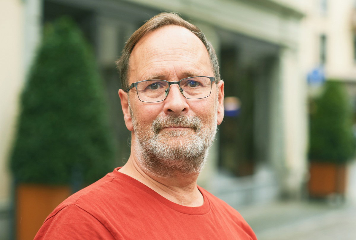 mężczyzna około 60 lat w czerwonej koszulce z zarostem i w okularach stoi na ulicy zatroskany