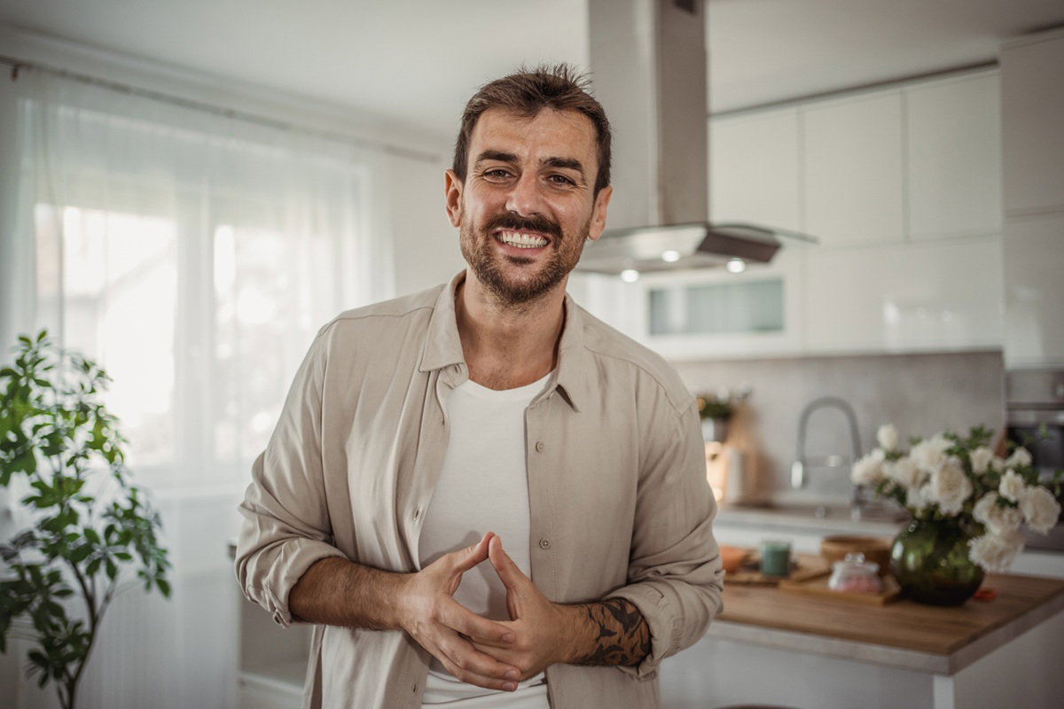 mężczyzna 35 plus z zarostem i w beżowej koszuli stoi w kuchni i się śmieje