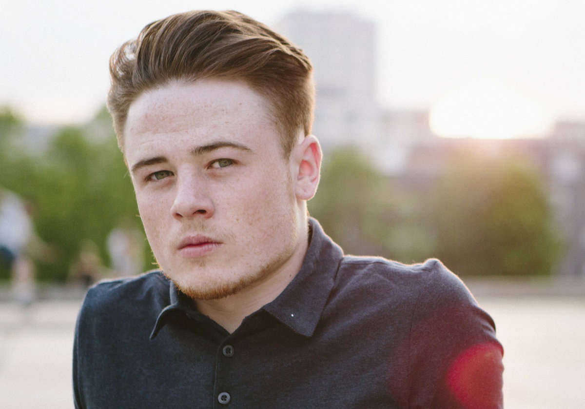 chłopak w wieku około 20 plus siedzi na zewnątrz na tle osiedla smutny i zamyślony