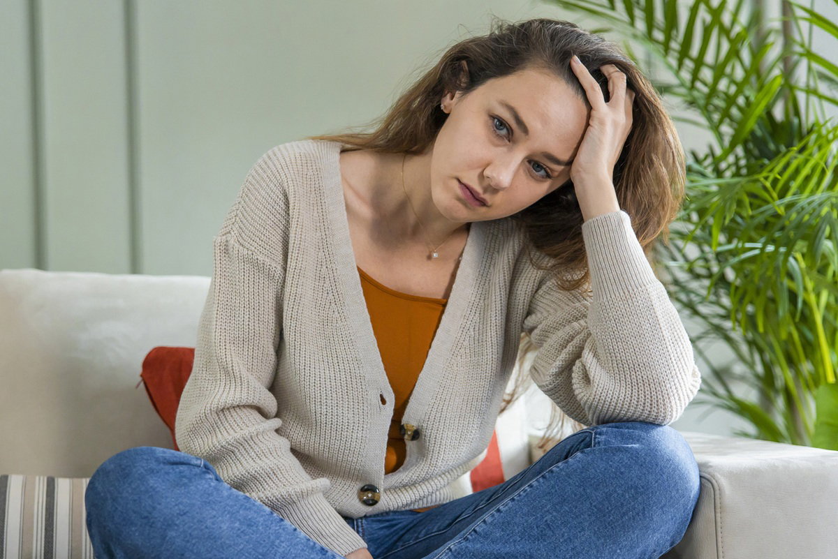 kobieta 30 plus siedzi na kanapie w beżowym swetrze i podpiera głowę ręką zmarrtwiona