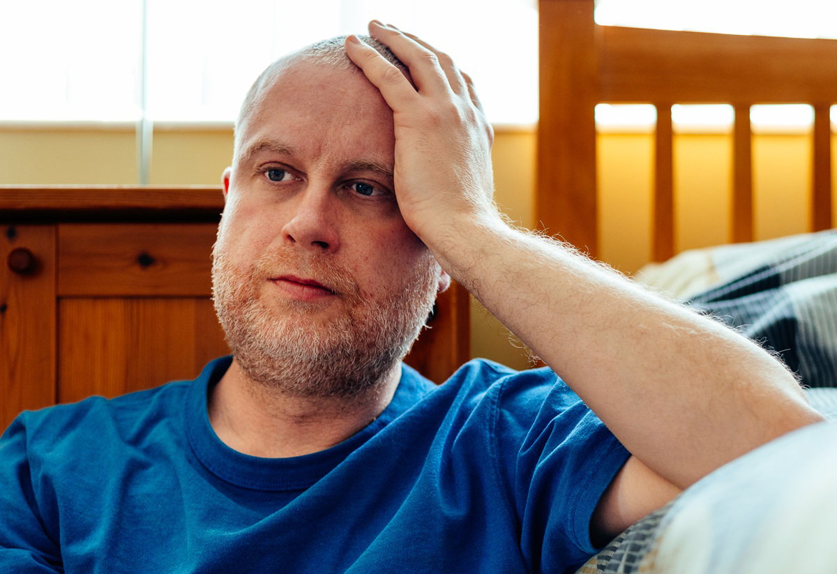 mężczyzna w wieku około 35 lat z zarostem w niebieskiej koszulce siedzi na podłodze przy łóżku załamany