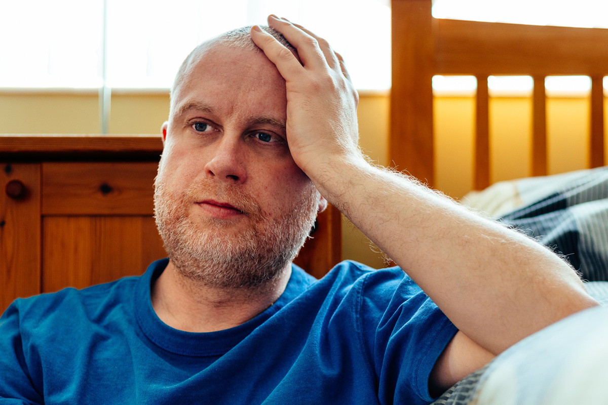 mężczyzna w wieku około 35 lat z zarostem w niebieskiej koszulce siedzi na podłodze przy łóżku załamany