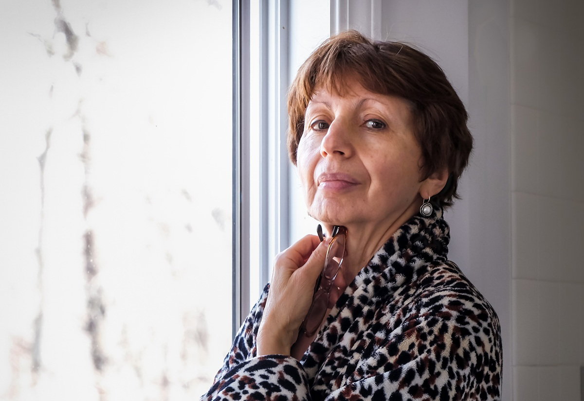 kobieta w wieku około 70 lat w szlafroku w panterkę stoi przy oknie zamyślona i smutna